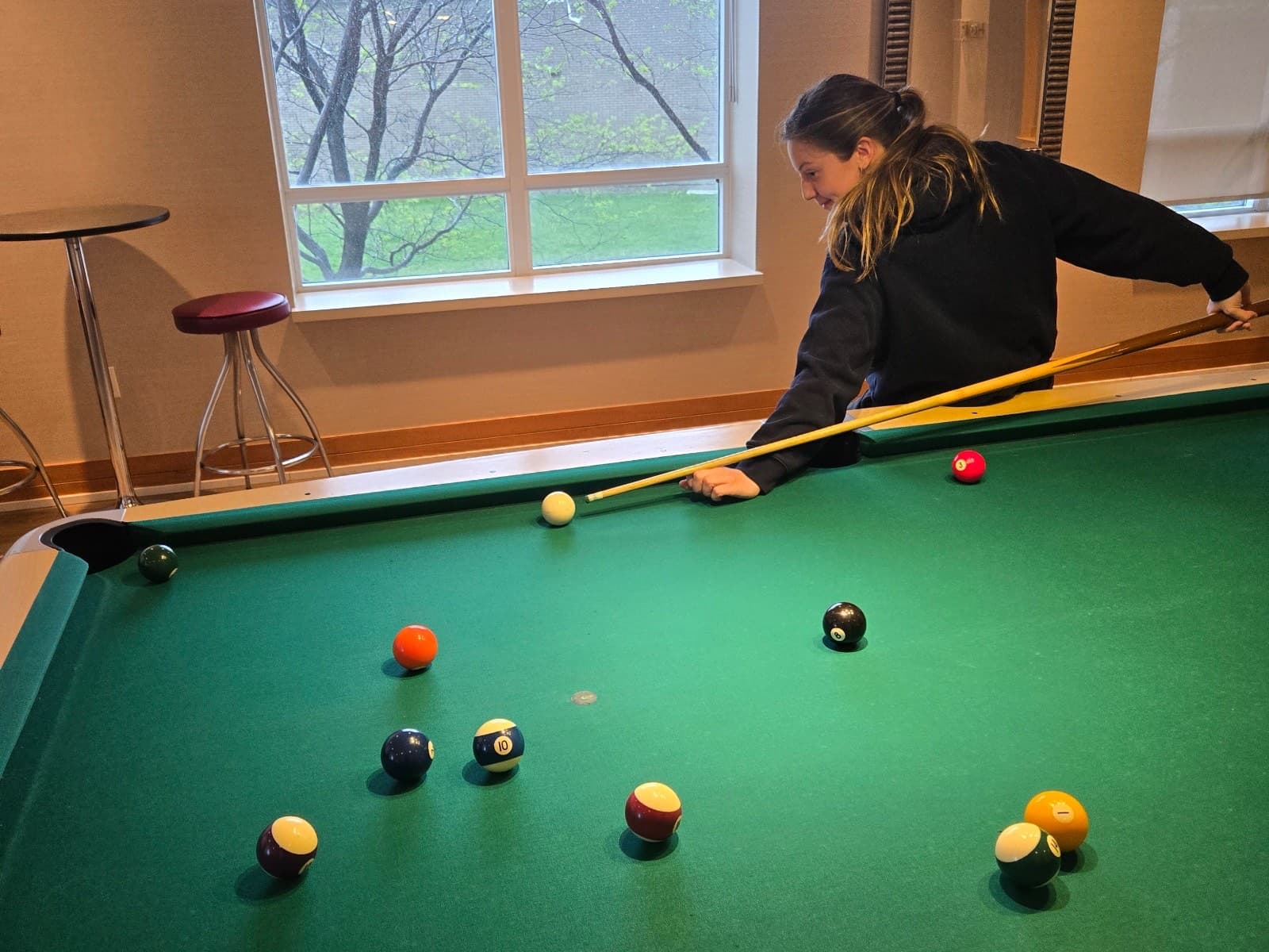 Chelsea leaning over a pool table, lining up a shot by a sunlit window